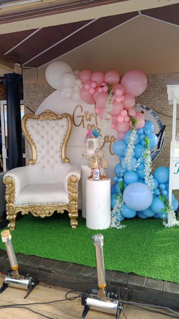 IMG-20250603-WA0022 Gender reveal display with white throne chair, pink and blue balloons, and dessert stands.
