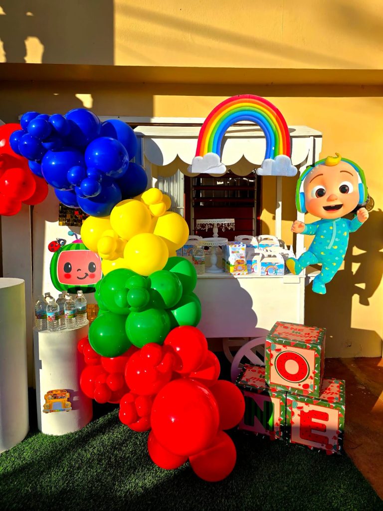 WhatsApp Image 2026-06-18 at 13.47.50 Rainbow-themed first birthday setup with colorful balloon column, character cutout, and treat cart.